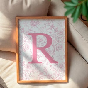 May include: A framed print featuring the letter "R" in a soft pink hue. The background showcases a delicate floral pattern in pink and white. The wooden frame adds a touch of warmth. The print rests on a beige cushion, with a plant in the background.