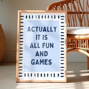 Puede incluir: Un cuadro enmarcado con el texto "ACTUALLY IT IS ALL FUN AND GAMES" se encuentra frente a una silla de ratán con un cojín blanco. La silla está en una habitación con plantas en macetas, creando un ambiente relajado y acogedor. El marco tiene un acabado de madera clara.