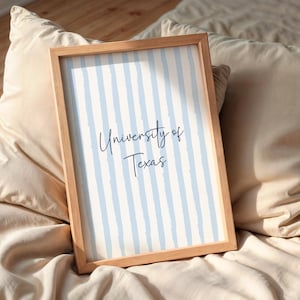 May include: A framed print with the text "University of Texas" in a handwritten style. The background has vertical light blue stripes on a white background. The frame is made of light-coloured wood. The print is resting on a bed with beige pillows.