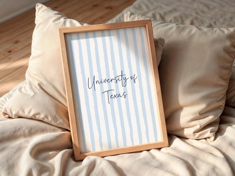 May include: A framed print with the text "University of Texas" in a handwritten style. The background has vertical light blue stripes on a white background. The frame is made of light-coloured wood. The print is resting on a bed with beige pillows.