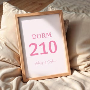 May include: A framed print with the word "DORM" above the number "210" in pink. Below the number are the names "Ashley & Sophia" in a script font. The frame is light brown, and the print rests on a bed with beige pillows.