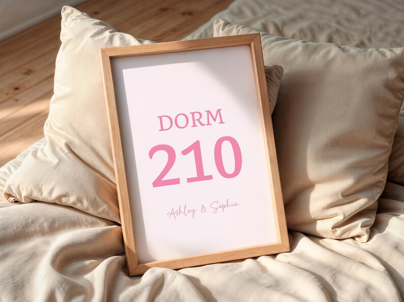 May include: A framed print with the word "DORM" above the number "210" in pink. Below the number are the names "Ashley & Sophia" in a script font. The frame is light brown, and the print rests on a bed with beige pillows.