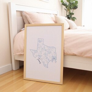 May include: A framed print of the state of Texas, with a light blue floral pattern inside the outline. The word "Texas" is written in cursive. The frame is light wood, and the print leans against a bed.