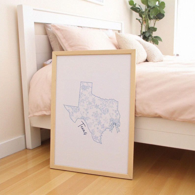 May include: A framed print of the state of Texas, with a light blue floral pattern inside the outline. The word "Texas" is written in cursive. The frame is light wood, and the print leans against a bed.