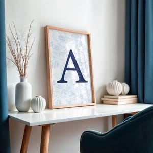 May include: A framed print with the letter "A" in navy blue, set against a floral pattern. The print sits on a white desk, alongside a vase of dried branches and decorative spheres. Teal curtains and a teal chair are visible.