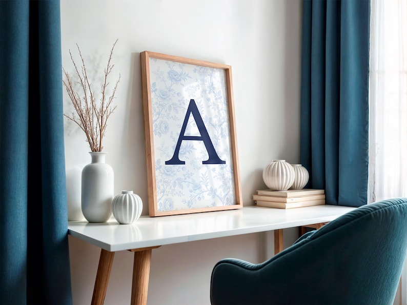 May include: A framed print with the letter "A" in navy blue, set against a floral pattern. The print sits on a white desk, alongside a vase of dried branches and decorative spheres. Teal curtains and a teal chair are visible.
