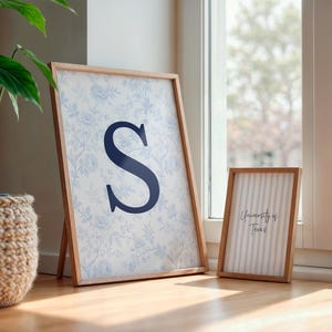 May include: Two framed prints with wooden frames. The larger print features a blue floral pattern and a navy blue letter "S". The smaller print has vertical blue stripes and the text "University of Texas". A potted plant is in the background.
