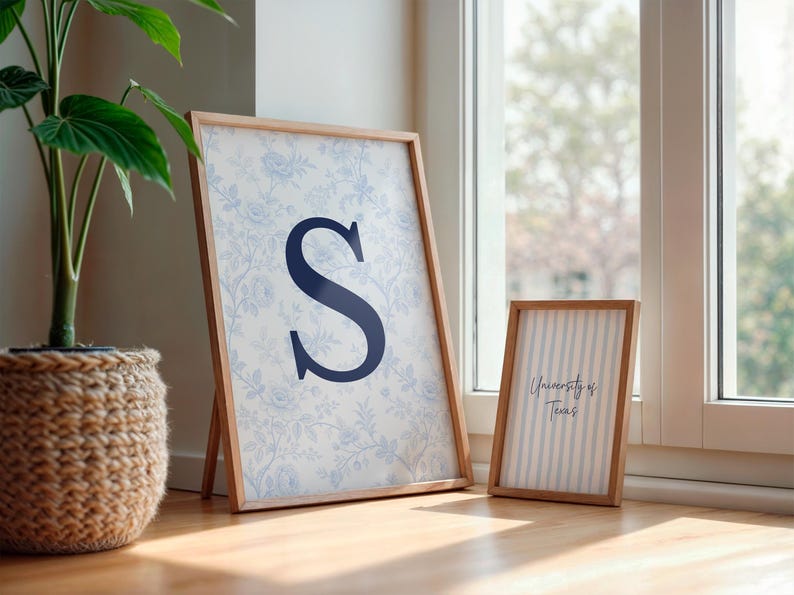 May include: Two framed prints with wooden frames. The larger print features a blue floral pattern and a navy blue letter "S". The smaller print has vertical blue stripes and the text "University of Texas". A potted plant is in the background.