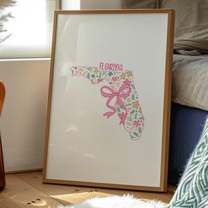 May include: A framed print featuring the outline of Florida filled with a floral pattern in pink, green, and yellow. The word "FLORIDA" is at the top, and a pink bow is at the bottom. The frame is made of light brown wood.