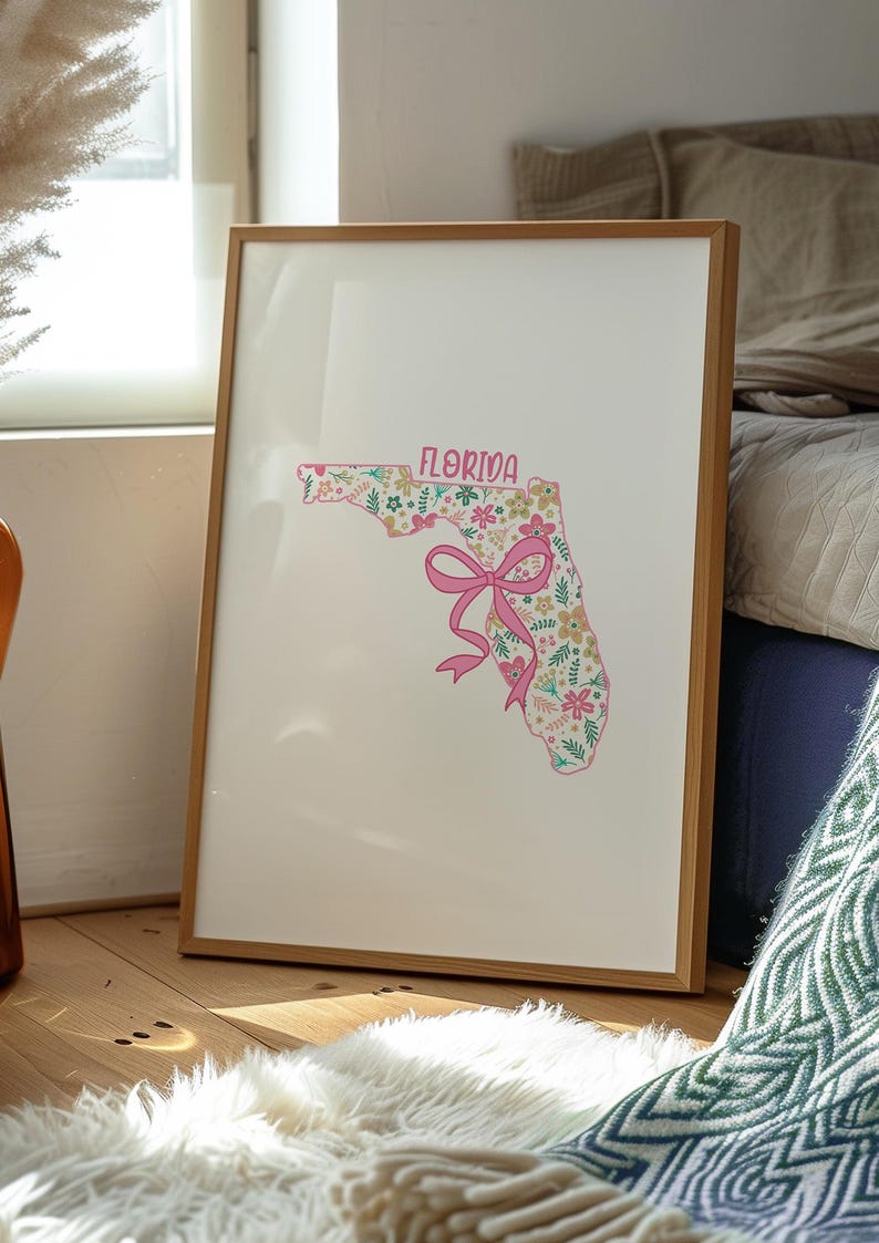 May include: A framed print featuring the outline of Florida filled with a floral pattern in pink, green, and yellow. The word "FLORIDA" is at the top, and a pink bow is at the bottom. The frame is made of light brown wood.
