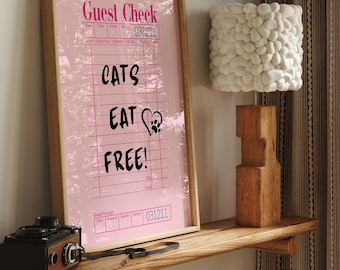 Könnte beinhalten: Gerahmter rosa "Guest Check" Druck mit dem Text "CATS EAT FREE!" und einem Pfotenabdruck-Herz. Eine Vintage-Kamera und eine Holzlampe mit weißem Steinschirm stehen auf einem Holzregal.