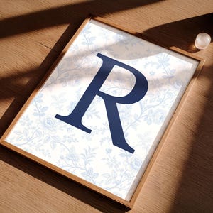 May include: A framed print with a navy blue letter "R" on a light blue floral background. The wooden frame adds a classic touch. The print is displayed on a wooden surface. The overall design is elegant and suitable for home decor.