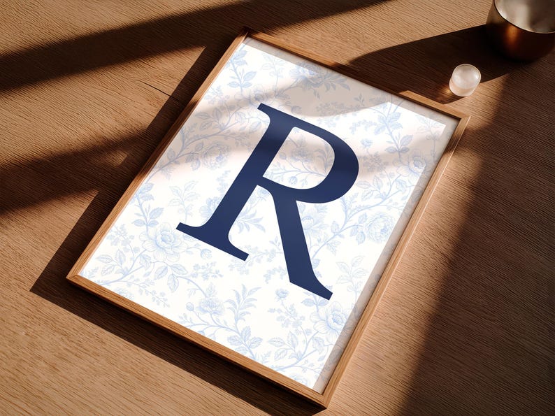 May include: A framed print with a navy blue letter "R" on a light blue floral background. The wooden frame adds a classic touch. The print is displayed on a wooden surface. The overall design is elegant and suitable for home decor.