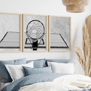 May include: A set of three black and white prints featuring a basketball hoop with a net. The prints are framed in a light wood frame and are hung on a white wall above a bed with blue bedding.