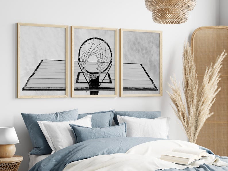 May include: A set of three black and white prints featuring a basketball hoop with a net. The prints are framed in a light wood frame and are hung on a white wall above a bed with blue bedding.