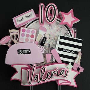 May include: A pink and white cake topper with a beauty theme. It features a pink makeup bag with the word "BEAUTY", a Sephora shopping bag, and a banner with the name "Valeria". The number "10" is also included.