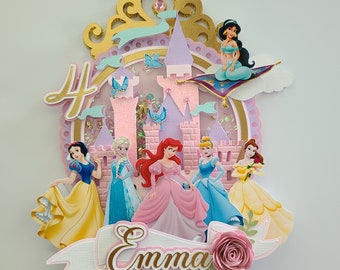 May include: A pink and gold cake topper featuring a castle, a golden crown, and seven Disney princesses. The princesses include Snow White, Ariel, Cinderella, Belle, Jasmine, Aurora, and Rapunzel. The cake topper also includes the number 4 and the name "Emma".