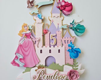 May include: A pink and purple castle cake topper with a Sleeping Beauty princess figure, a gold number 5, and three fairies. The cake topper has a white banner with the name Penelope written in gold.