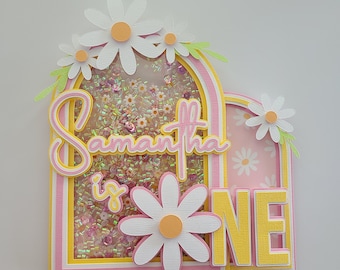 May include: A pink and yellow cake topper with the name Samantha and the words "is one" in a whimsical font. The topper features a glittery background and several white daisies.