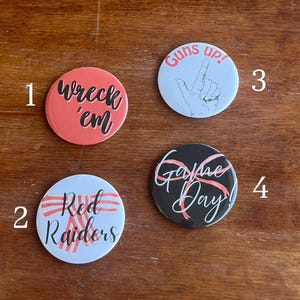May include: Four round buttons with text. One is coral with "wreck 'em" in black script. Another is white with "Red Raiders" in red script. A third is white with "Guns up!" and a hand gesture. The last is black with "Game Day!"