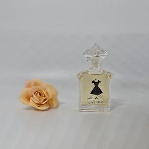 May include: A clear perfume bottle with a decorative stopper, featuring a black dress illustration and script. A peach-coloured rose lies beside the bottle. The perfume bottle is filled with a light amber liquid.
