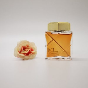 以下が含まれることがあります： 金色のキャップと琥珀色の液体が入った正方形の香水瓶。「KL HOMME EAU DE TOILETTE」と書かれています。ボトルの左側には、薄いピンクと赤のバラがあります。背景は白です。