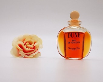 May include: A small, clear glass bottle of Dune Eau de Toilette by Christian Dior Paris. The bottle is filled with a golden-amber liquid and has a gold cap. A small, yellow and red rose is in the background.
