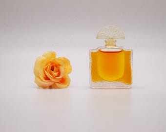 May include: A clear glass bottle of perfume with a yellow liquid and a white, ornate stopper. A single, light orange silk flower is in the foreground.