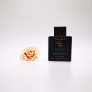以下が含まれることがあります： 黒い長方形の香水瓶。金色の文字で「Van Cleef & Arpels Paris pour homme」と書かれています。7mlのオードトワレ入り。左側に、赤いアクセントのある、桃色のバラがあります。