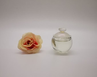 May include: A clear, round glass jar with a lid, next to a peach-colored rose. The jar has a small, round lid and is filled with a clear liquid. The rose has a soft, layered appearance with red and yellow hues.