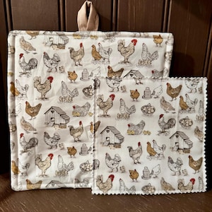 Puede incluir: Juego de manopla y agarradera blancos con un estampado repetido de gallinas, gallineros y huevos. La manopla tiene un lazo beige para colgar. La agarradera tiene un borde festoneado.