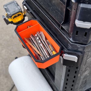 May include: An orange and black tool organizer with a magnetic latch, attached to a black toolbox. The organizer is filled with various tools, including drill bits and a pen.