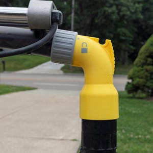 May include: A yellow plastic elbow connector attached to a black pole with a grey locking collar. The connector features a printed padlock icon and a small square symbol, indicating a secure locking mechanism for the attached equipment components.