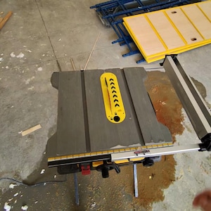 Może przedstawiać: Szara piła stołowa z żółtą osłoną i srebrną szyną prowadzącą. Piła ma czarną podstawę i stoi na betonowej podłodze. Dodatkowe żółte i niebieskie półki znajdują się w tle.