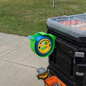 Może przedstawiać: Zielony, niebieski i żółty dozownik taśmy drukowany w 3D, zamontowany na czarnej skrzynce narzędziowej. Dozownik posiada mechanizm blokujący i obracający uchwyt.