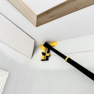 May include: A yellow and black corner measuring tool is positioned in the corner of a room. The tool is used for measuring angles and distances. The tool is attached to a black pole.
