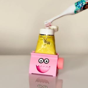 Op de afbeelding: Een roze tandenborstelhouder met een cartoongezicht en een gele tube tandpasta. Een witte en blauwe elektrische tandenborstel is boven de tube tandpasta geplaatst. De houder heeft een lachend gezichtsontwerp.