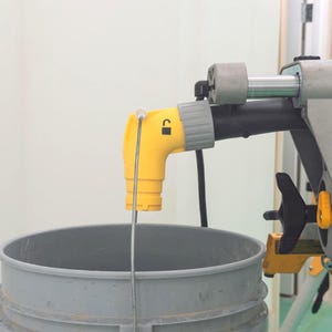 May include: A yellow and grey paint sprayer nozzle is positioned over a grey bucket. The nozzle has a lock symbol and a flexible hose. The sprayer is part of a larger painting tool, with a black handle and grey components.