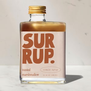 May include: A square glass bottle of toasted marshmallow simple syrup. The label features the word "SURRUP" in large, brown letters. The bottle is filled with a light brown liquid and topped with a gold cap. The bottle contains 11.16 oz (330 mL) of syrup.