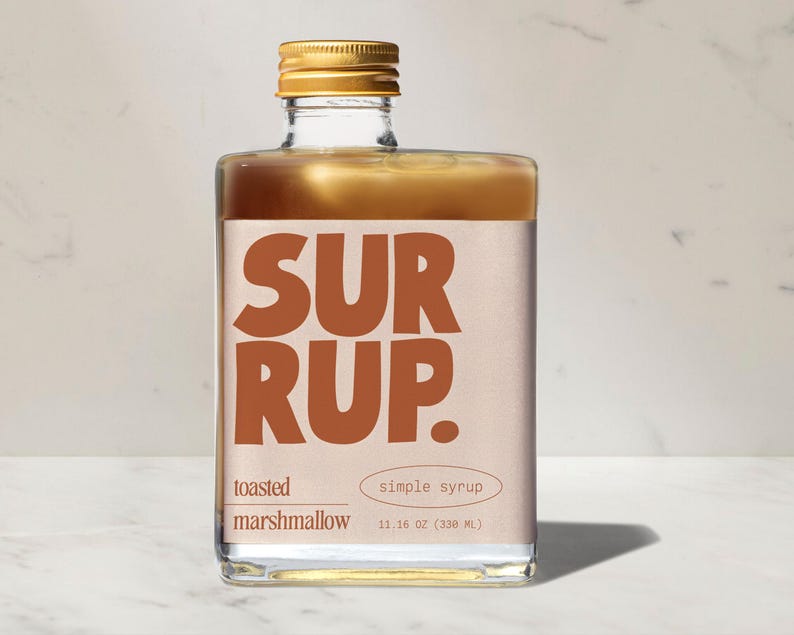 May include: A square glass bottle of toasted marshmallow simple syrup. The label features the word "SURRUP" in large, brown letters. The bottle is filled with a light brown liquid and topped with a gold cap. The bottle contains 11.16 oz (330 mL) of syrup.