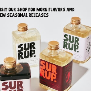 May include: Four square glass bottles of syrup with gold caps are displayed. Each bottle has a label with the word "SUR RUP" in bold letters. The text "VISIT OUR SHOP FOR MORE FLAVORS AND NEW SEASONAL RELEASES" is at the top.