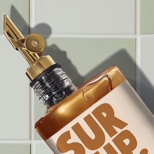 May include: A gold-colored bottle with a matching spout and a black rubber seal. The bottle has a rectangular shape and the text "SUR UP" in a brown color. The background is a light green and white tiled wall.