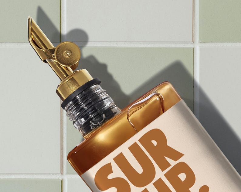 May include: A gold-colored bottle with a matching spout and a black rubber seal. The bottle has a rectangular shape and the text "SUR UP" in a brown color. The background is a light green and white tiled wall.