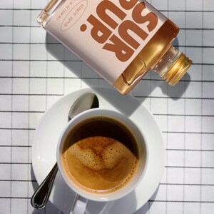 May include: A white coffee cup and saucer with a spoon, next to a bottle of toasted marshmallow simple syrup. The syrup bottle is labeled "SUR RUP" and "Simple Syrup" with a 11.16 fl oz (330 ml) measurement.