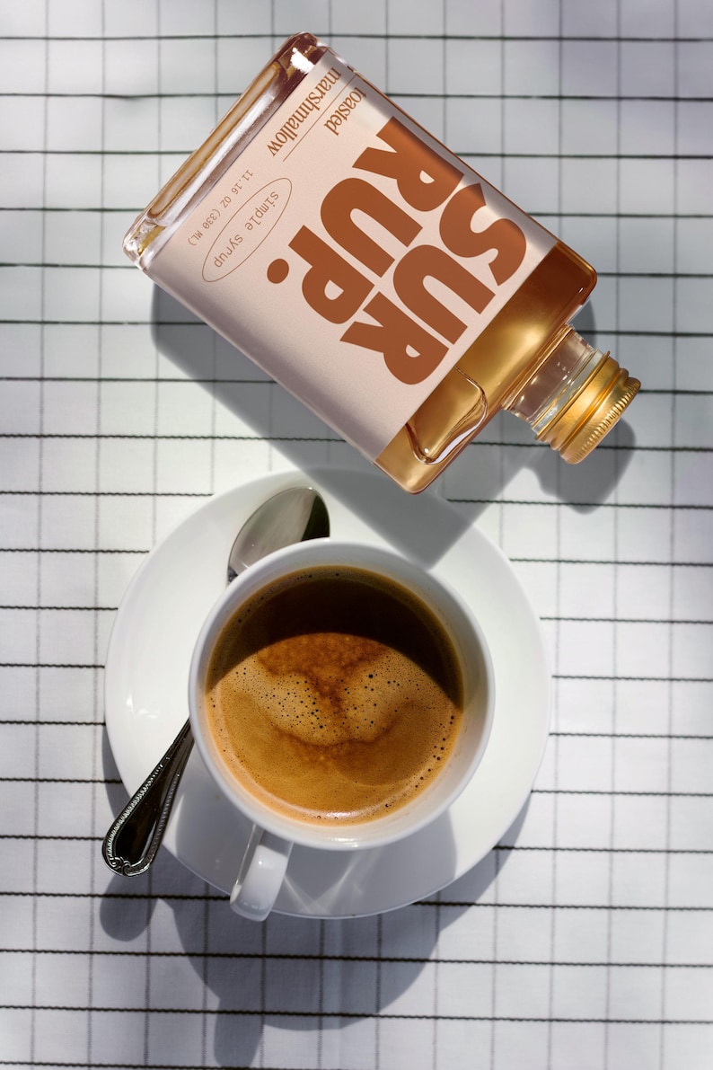 May include: A white coffee cup and saucer with a spoon, next to a bottle of toasted marshmallow simple syrup. The syrup bottle is labeled "SUR RUP" and "Simple Syrup" with a 11.16 fl oz (330 ml) measurement.