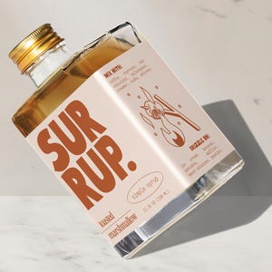 May include: A square glass bottle of toasted marshmallow simple syrup with a gold cap. The label reads "SUR RUP." and "simple syrup." It suggests mixing with coffee, chocolate, and drizzling on ice cream and pancakes. The bottle contains 11.16 oz (330 ml).