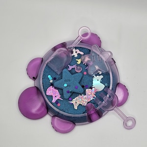 May include: A purple plastic sand tray with a blue sand base and various unicorn and star shaped toys. The tray has a handle and a scoop.