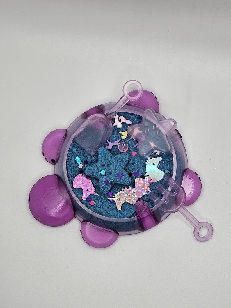 May include: A purple plastic sand tray with a blue sand base and various unicorn and star shaped toys. The tray has a handle and a scoop.