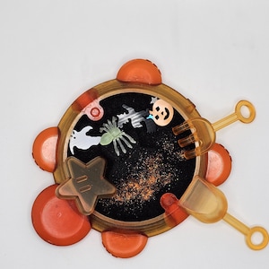 May include: Orange and black Halloween themed sensory toy with a black sand-like material inside. The toy has a star, spider, ghost, and pumpkin shapes inside. It comes with a small shovel and a rake.