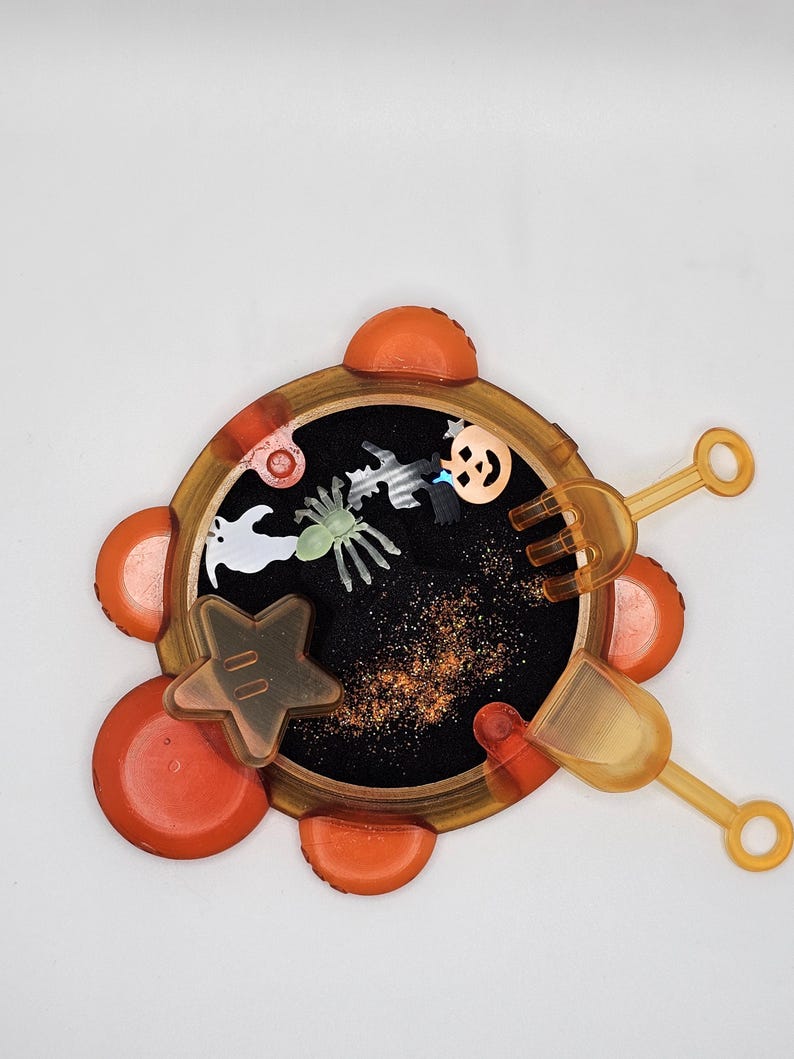 May include: Orange and black Halloween themed sensory toy with a black sand-like material inside. The toy has a star, spider, ghost, and pumpkin shapes inside. It comes with a small shovel and a rake.